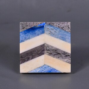 Könnte beinhalten: Eine quadratische Dekorplatte mit einem Chevron-Muster. Die Platte besteht aus Steinen in verschiedenen Farben, darunter Blau, Weiß, Schwarz und Beige.