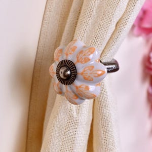May include: A flower-shaped curtain tieback featuring a white ceramic base with orange leaf patterns. The center of the flower has a dark metallic circular accent with a ridged edge, and the hardware is attached to a beige textured fabric curtain.