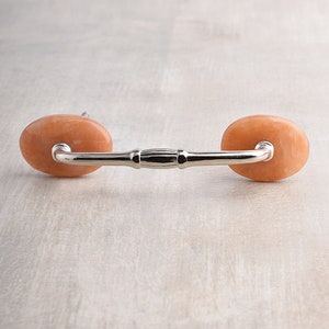 May include: A cabinet handle featuring a polished silver-toned metal bar connected to two smooth, oval-shaped peach-colored stone accents on each end. The hardware has a central decorative detail on the bar and a clean, symmetrical design.