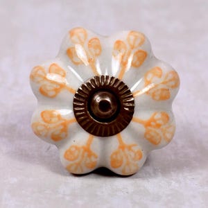 Peut inclure: Un bouton de meuble en céramique en forme de fleur avec une base blanche et des motifs de feuilles orange. Le centre présente un élément métallique circulaire et nervuré de couleur bronze foncé servant de point de fixation au bouton.