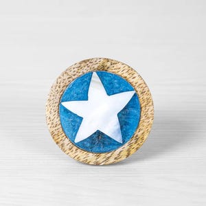 以下が含まれることがあります： 青と白の星形のデザインが施された丸い木製ノブ。星は真珠貝で作られており、青い円の中にセットされています。ノブは木製のフレームで囲まれています。
