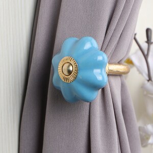 May include: A light blue ceramic curtain tieback with a gold metal ring. The tieback is shaped like a flower with a raised center.