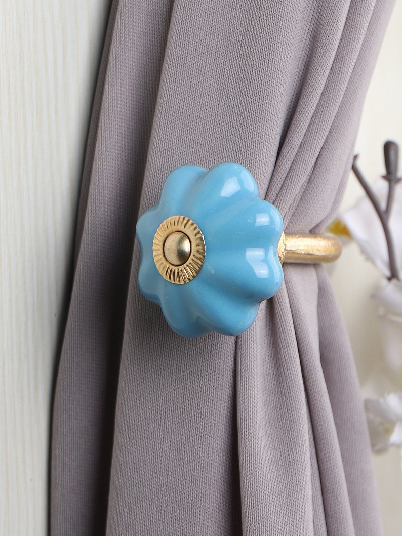 May include: A light blue ceramic curtain tieback with a gold metal ring. The tieback is shaped like a flower with a raised center.