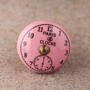 Peut inclure: Bouton de porte en céramique rose avec un design de cadran d'horloge. Le cadran de l'horloge a des chiffres et des aiguilles noirs, ainsi que le texte "PARIS CLOCKS" en noir.