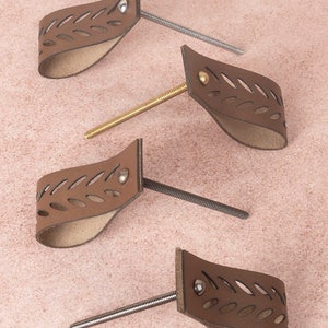 Könnte beinhalten: Vier braune Leder-Schrankgriffe mit einem Ausschnittmuster. Jeder Griff hat eine Schraube aus Metall und einen Niet aus Metall.