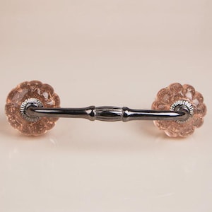 May include: A drawer pull featuring a dark metallic handle connected to two round, fluted, light pink glass knobs. The glass knobs have a textured, speckled appearance, and the metal hardware has a smooth, polished, dark grey finish.