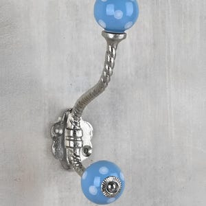 May include: A decorative wall hook with a silver finish. The hook features two light blue, polka-dotted ceramic spheres. The curved metal arm has a twisted design. The hook is mounted on a decorative silver base.