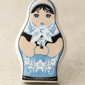 May include: A ceramic doll figurine with a blue and white dress, black hair, and a blue headscarf. The doll is holding a bouquet of flowers.