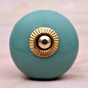 May include: A round, matte teal ceramic cabinet knob featuring a central gold-toned metal accent. The gold hardware has a circular, fluted, or ribbed design surrounding a smooth, raised centre point, providing a contrast to the solid teal surface.