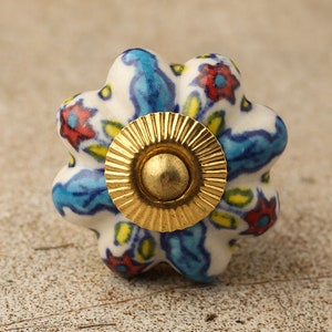 Op de afbeelding: Een witte keramische knop met een blauw, rood en geel bloemenpatroon. De knop heeft een gouden metalen basis.