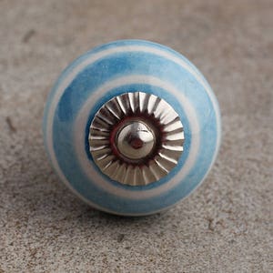 Puede incluir: Un pomo de cerámica rayado azul y blanco con una base de metal plateado. El pomo es redondo y tiene un acabado liso.