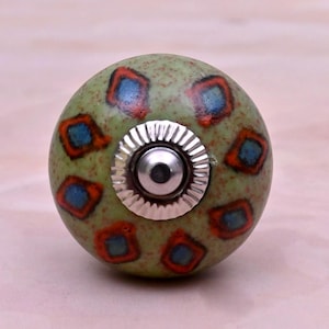 Op de afbeelding: Een ronde keramische meubelknop met een gespikkelde lichtgroene basis. Het oppervlak heeft een herhalend patroon van blauwe ruiten met oranje omlijningen. Een zilverkleurig metalen beslag met een geribbelde rand is in het midden geplaatst.