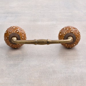 May include: A brown ceramic drawer pull with a gold metal bar connecting two round knobs. The knobs have a white floral design.
