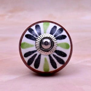 Op de afbeelding: Een ronde keramische meubelknop met een witte basis en een radiaal patroon van zwarte en limoengroene bloemblaadjes. De knop heeft een donkerbruine buitenrand en een centraal zilverkleurig metalen onderdeel met een geribbeld ontwerp.