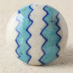 May include: A round, ceramic cabinet knob with a white base and a turquoise zigzag pattern. The knob has a glossy finish and is likely used for furniture or decorative purposes.