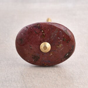 Könnte beinhalten: Ein ovaler Knauf aus Naturstein mit einem gesprenkelten rot-braunen Muster. Der Knauf hat eine goldene Schraube.