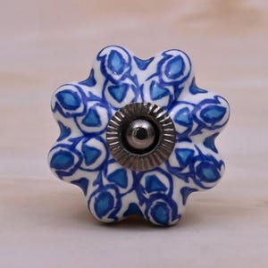 May include: A flower-shaped ceramic cabinet knob featuring a white base with a repeating blue and light blue abstract pattern. The centre of the knob has a circular, ridged metallic silver-toned hardware component for mounting purposes.