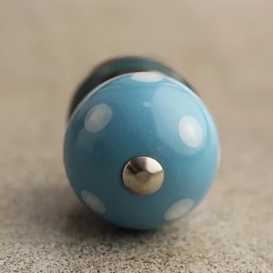 Puede incluir: Un pomo de cerámica azul con lunares blancos y un tornillo plateado. El pomo es redondo y tiene una superficie lisa.