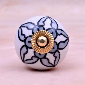 Op de afbeelding: Een ronde witte keramische meubelknop met een handgeschilderd lichtblauw bloemmotief omlijnd met zwarte inkt. Het midden van de knop is voorzien van een cirkelvormig, geribbeld goudkleurig metalen beslag met een gladde, bolvormige bovenkant.