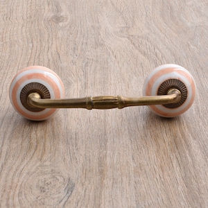 Può includere: Una maniglia per cassetto in ottone con due pomelli rotondi in ceramica. I pomelli sono bianchi con strisce rosa e marroni.