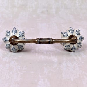May include: A drawer pull featuring two flower-shaped ceramic knobs with a white base and blue leaf pattern. The knobs are connected by a horizontal, antique-finished brass metal handle with a central decorative band and fluted metal collars.