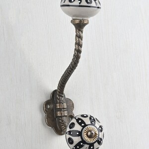 May include: A wall-mounted hook featuring a curved, textured bronze-toned metal arm and two ceramic knobs. The knobs are white with black painted floral patterns and small white dots. The base plate has a scalloped edge with two screw holes.