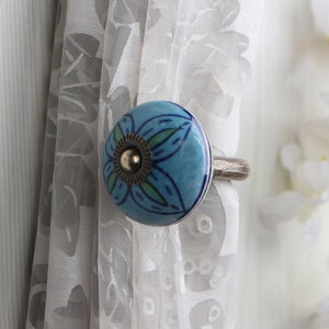 May include: A blue and green ceramic curtain tieback with a silver metal ring. The tieback features a floral design.