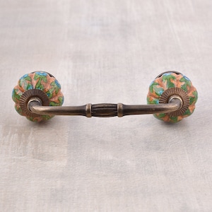 May include: A pair of ceramic drawer pulls with a floral design in shades of green, blue, and orange. The pulls are attached to a metal bar with a decorative ribbed design.