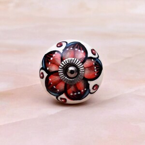 May include: A round ceramic cabinet knob featuring a hand-painted floral design with red and black petals on a white background. The center of the knob has a circular silver-toned metal hardware piece with a ridged edge and a central screw hole.