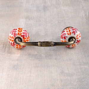 Può includere: Una maniglia per cassetto in ceramica con una barra in ottone e due pomelli rotondi. I pomelli sono bianchi con un motivo floreale rosso e arancione.
