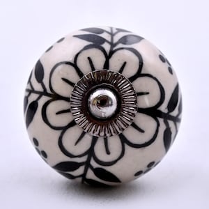 May include: A round ceramic cabinet knob featuring a cream-colored base with a hand-painted black floral and leaf pattern. The center of the knob is accented with a circular, ridged silver-toned metal cap and a small central screw head.