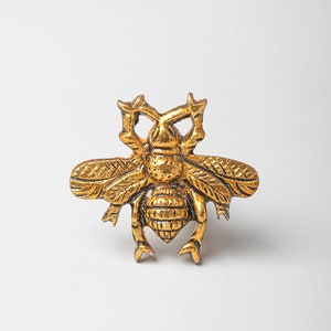 Może przedstawiać: Złoty, ozdobny przedmiot w kształcie pszczoły z misternymi detalami. Pszczoła ma fakturowane ciało i skrzydła.