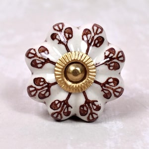 Peut inclure: Un bouton de meuble en céramique blanche en forme de fleur festonnée, orné de motifs de feuilles peints en brun. Le centre du bouton présente une pièce métallique dorée circulaire avec un bord strié et un dôme central lisse et arrondi.