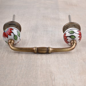 May include: Brass cabinet pull with ceramic knobs. The knobs are white with red flowers and green leaves.