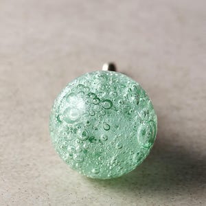 May include: A round, light green glass knob with a bubbly texture. The knob has a silver metal stem. The glass is translucent, and the bubbles are of varying sizes. This decorative knob could be used for furniture or other DIY projects.