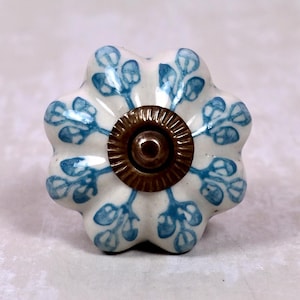 Peut inclure: Un bouton de meuble en céramique en forme de fleur avec une base blanche et des motifs de feuilles peints en bleu. Le centre du bouton présente un élément matériel métallique de couleur bronze, circulaire et nervuré, avec un petit trou.