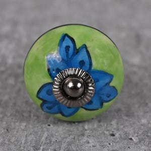 Puede incluir: Pomo de armario redondo de cerámica con un diseño pintado a mano. El pomo presenta una base verde brillante con un diseño floral azul. Los herrajes metálicos son de color plata oscura.