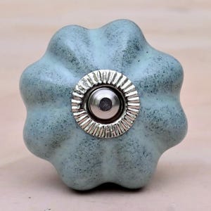 Puede incluir: Un tirador de cerámica para muebles con forma de calabaza en color verde salvia polvoriento con acabado moteado. El centro presenta una placa metálica circular plateada con borde estriado y un botón plateado liso y elevado.