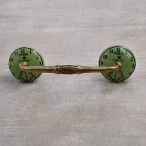 以下が含まれることがあります： 緑色のセラミック製の引き出しの取っ手。時計の文字盤のデザインで、文字盤には「Paris Clocks」と黒色の数字と針が書かれています。取っ手は真鍮の棒に取り付けられています。