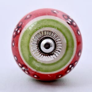 Könnte beinhalten: Ein runder Keramik-Möbelknopf mit roter Basis und einem hellgrünen Kreisband in der Mitte. Die Oberfläche ist mit kleinen weißen und schwarzen Blumenmustern verziert. Ein silberfarbenes Metallbeschlagteil mit geriffeltem Rand ist mittig.