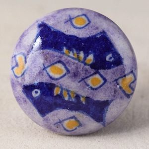 May include: A round ceramic knob with a purple background and a blue and yellow fish design.