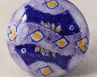 May include: A round ceramic knob with a purple background and a blue and yellow fish design.