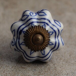 Puede incluir: Un pomo de cajón de cerámica blanca con forma festoneada similar a una flor, que presenta patrones de líneas azules pintadas a mano. El centro del pomo está acentuado con una pieza de metal circular y dentada en tono latón antiguo.