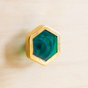 Op de afbeelding: Een zeshoekige kastknop met een gouden metalen frame en een groene glazen inzet. Het glas heeft een draaiende structuur.