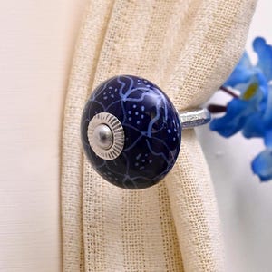 May include: A round, dark blue ceramic curtain tieback featuring a white hand-painted floral pattern and small dots. The knob has a silver-toned metal center cap with a ridged edge and a matching silver-toned metal post extending from the back.