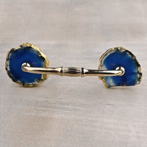 Peut inclure: Une poignée de porte en métal doré avec des extrémités en agate bleue. La poignée a une forme arrondie et lisse et une finition polie.