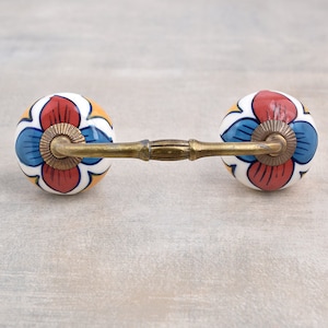 May include: A brass drawer pull with two ceramic knobs. The knobs are round and feature a floral design in shades of red, blue, yellow, and white.