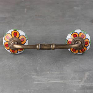 Può includere: Una maniglia per cassetto in ottone con due pomelli in ceramica. I pomelli sono bianchi con un motivo floreale in arancio, rosso e verde.