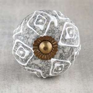 Könnte beinhalten: Ein grauer Keramik-Knopf mit einem weißen geometrischen Muster und einer goldenen Metallbasis.