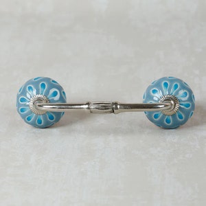 Puede incluir: Un par de tiradores de cajón de cerámica azul con un diseño floral blanco. Los tiradores están conectados por una barra de metal plateado.